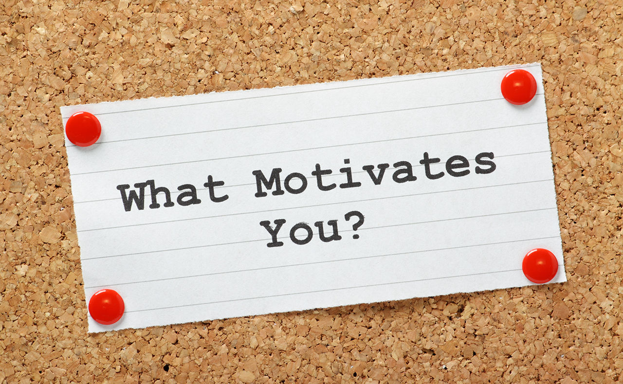 Piece of paper printed with "What Motivates You?" is thumb-tacked to a cork board.